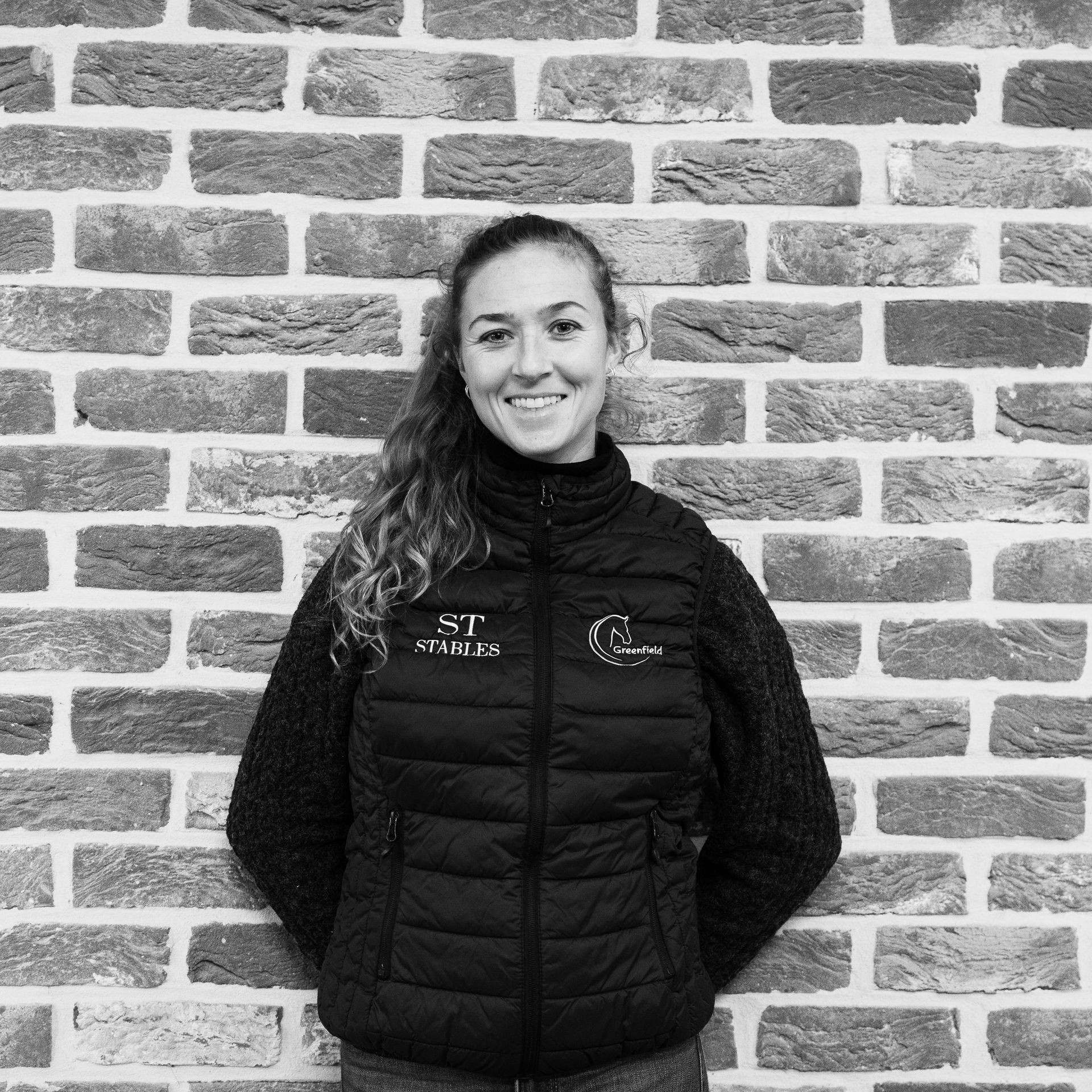 Person smiling in a black vest standing against a brick wall backdrop.